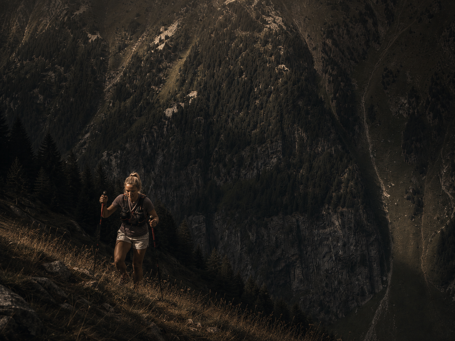 Female endurance athlete climbing a steep mountain trail with trekking poles.