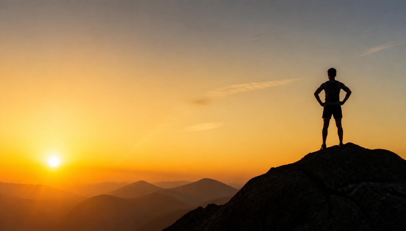 Runner standing on a mountain summit silhouetted against a golden sunrise.